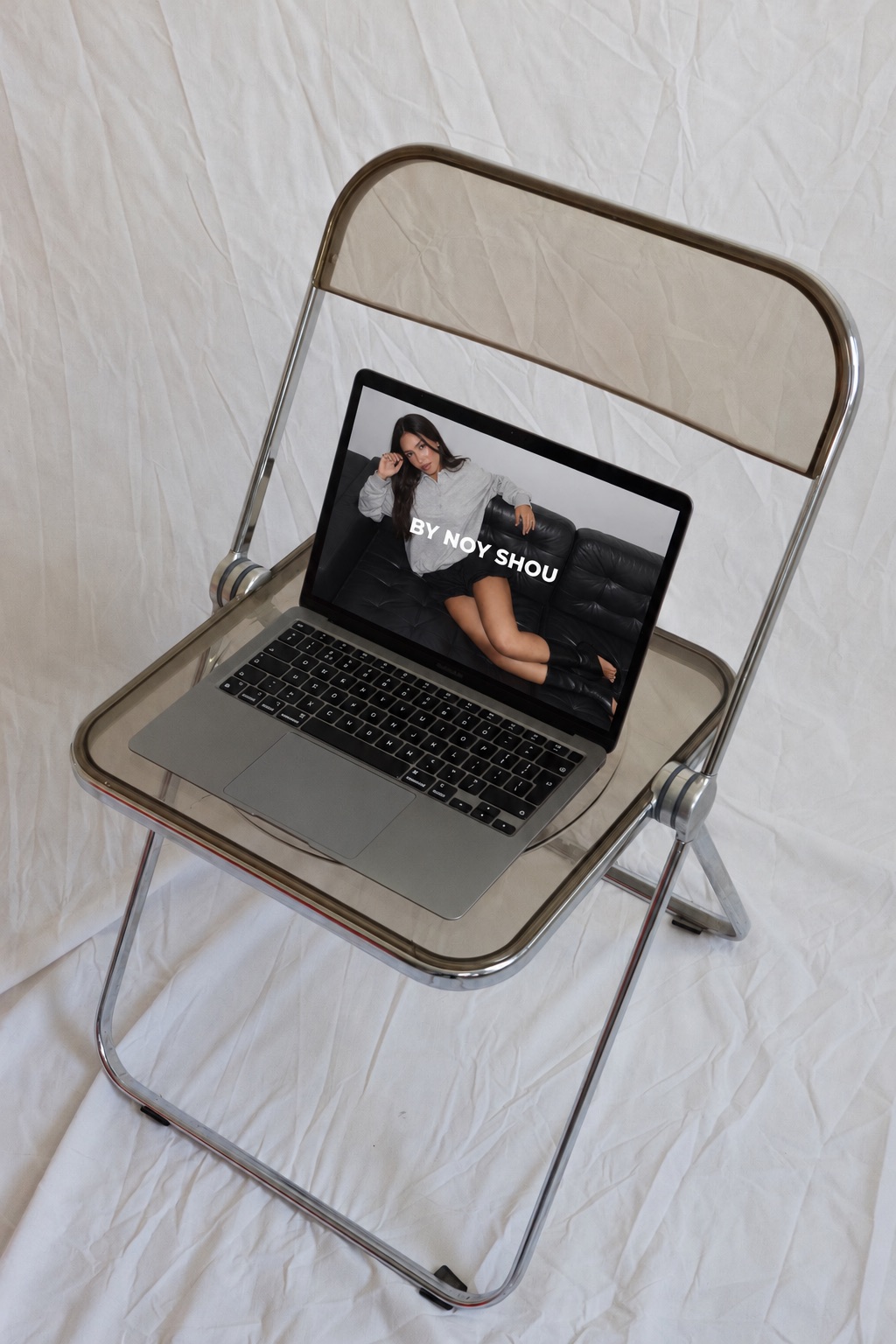 The BY NOY SHOU website on a laptop resting on a clear folding chair