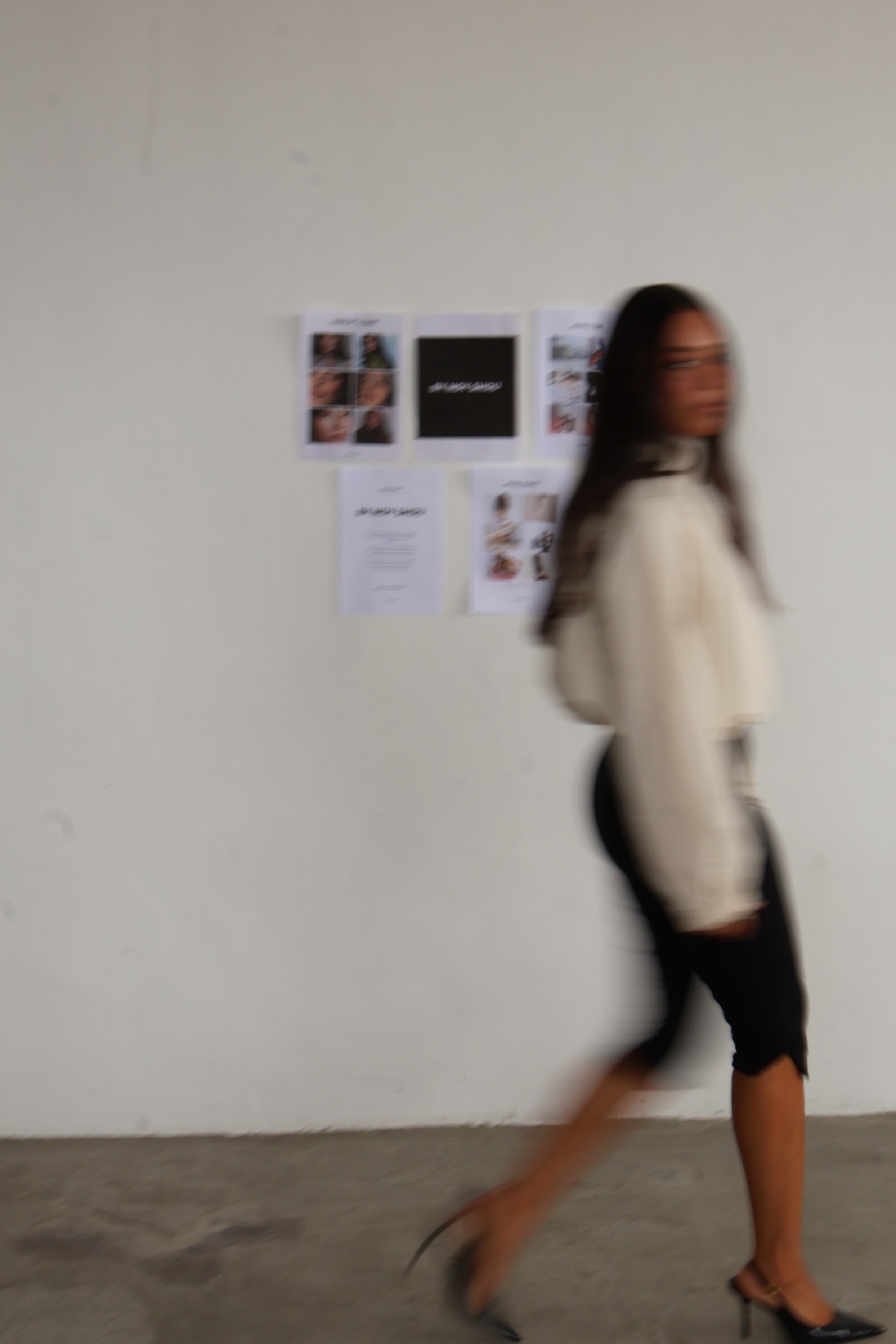 Noy walking past BY NOY SHOU posters on a white wall