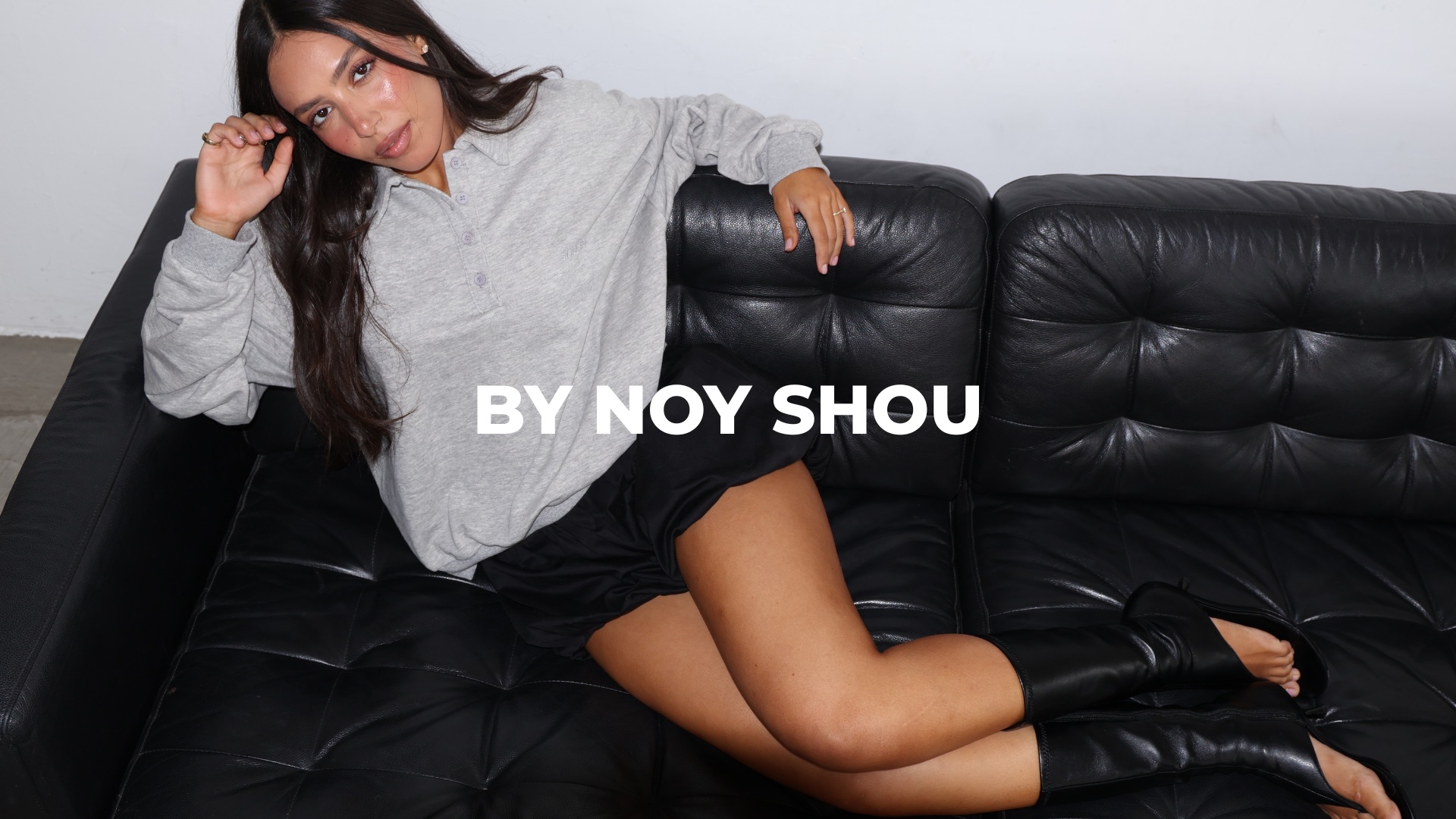 Noy Shou in a grey polo sweatshirt on a black leather sofa — BY NOY SHOU